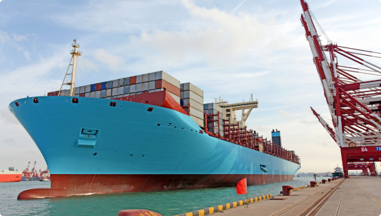 Большой бирюзовый контейнеровоз, груженый контейнерами, пришвартован у причала в порту под голубым небом с облаками, видны портовые краны красного и белого цвета